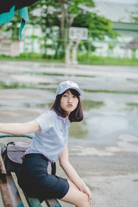 Portrait of smiling young woman sitting on bench in park during rainy season