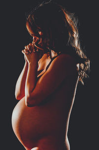 Side view of naked pregnant woman praying against black background