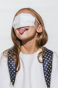 Playful girl sticking out tongue while covering eyes with protective face mask during covid-19