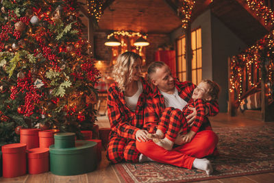 Smile active parents with small son in red checkered sleepwears waiting santa indoor.