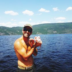 Portrait of shirtless man holding fish while standing in sea against sky