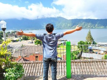 Rear view of young man with arms outstretched standing against houses and lake