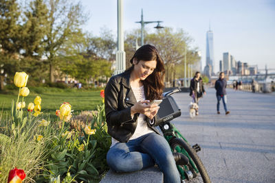Woman using smart phone while sitting on retaining wall with one world trade center in background