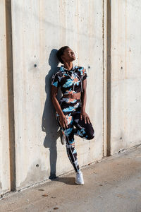 Cheerful confident african american female athlete in flowered sport clothes and white sneakers with closed eyes with interest and laughing while standing alone leaning on concrete wall in sunbeams and resting after training in city
