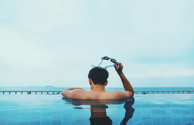 Rear view of shirtless man holding swimming goggles in swimming pool against sky