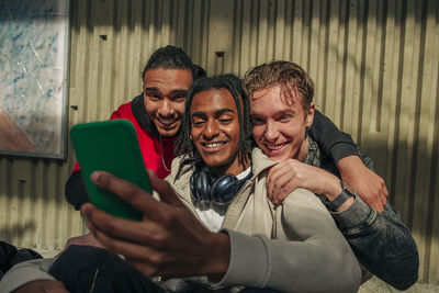 Smiling multiracial male friends doing video call through smart phone against wall
