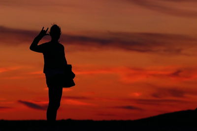 Silhouette woman gesturing horn sign while standing against orange sky