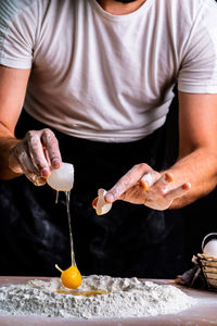 Faceless female baker breaking raw chicken egg into sifted flour pile on table cooking bread  person