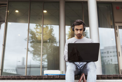 Low angle view of university student wearing headphones while using laptop against glass window at campus