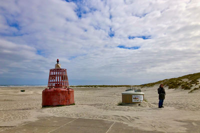 Beach under a blue and clouded sky with on the sand a red buoy. a waste container and a person