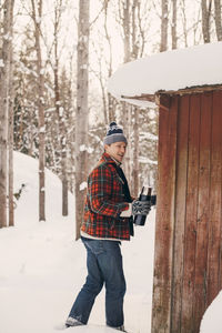 Smiling man holding drink container while walking by log cabin on snowy landscape