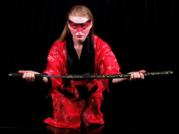 Portrait of young female model holding samurai sword while kneeling against black curtain