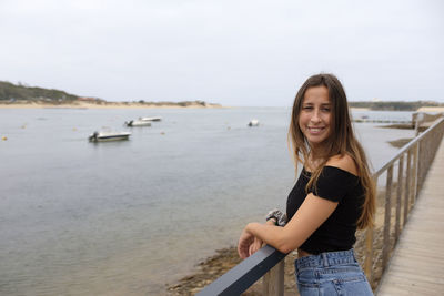 Portrait of smiling young woman standing by railing at beach