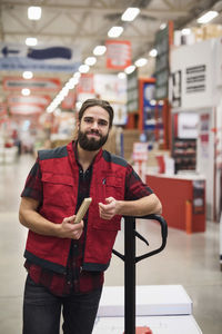 Portrait of confident salesman holding folding ruler while standing by handtruck in hardware store