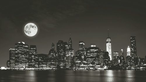 Illuminated cityscape by river against moon