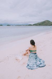 Rear view of woman relaxing on sand at beach against cloudy sky