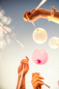 Low angle view of hands holding helium balloons against sky