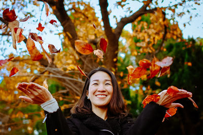 Smiling mid adult woman throwing autumn leaves while standing against trees in park