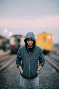 Portrait of man wearing hooded jacket while standing against trains at railroad tracks