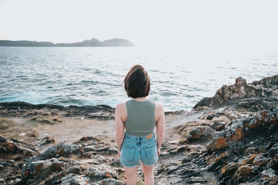 Rear view of woman standing on rock by sea against clear sky