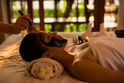 Cropped hand applying facial mask to young woman lying on bed in spa