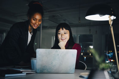 Smiling female entrepreneurs discussing while looking at laptop in creative office working late