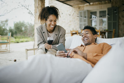 Cheerful man lying on sofa sharing smart phone with friend at backyard