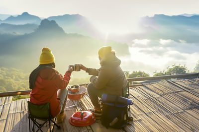 Friends having drink while sitting against mountains