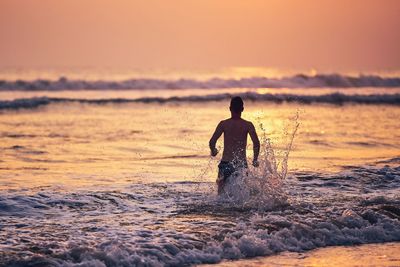 Man splashing water while running in sea against sky during sunset