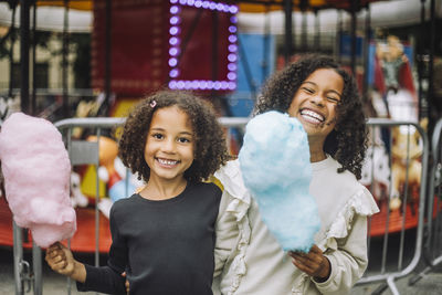 Portrait of cheerful sisters holding cotton candies while standing in front of carousel at amusement park