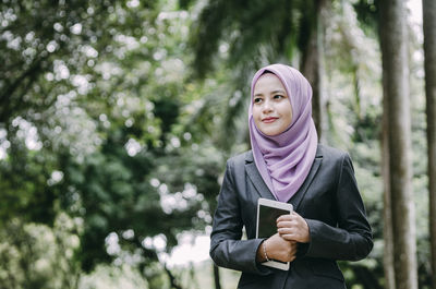 Businesswoman in hijab holding digital tablet against trees