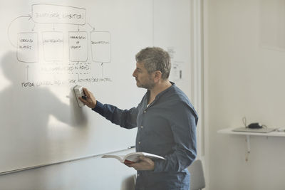 Side view of male teacher erasing diagram from whiteboard while holding book in classroom