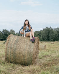 Full length of woman with dog sitting on haystack at field
