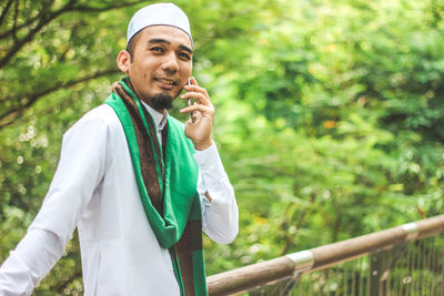 Smiling man in traditional clothing talking on mobile phone standing against trees