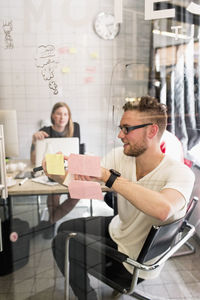 Young businessman writing ideas on adhesive notes with female colleague in background at new office
