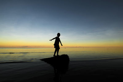 Silhouette girl standing on fallen tree in sea against sky during sunset