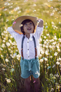 Funny happy child sit in hat on field with white dandelions  sunset in summer. soap bubbles  flying