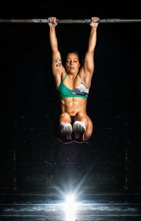 Full body muscular female in sportswear hanging on bar and lifting legs during functional workout in dark gym