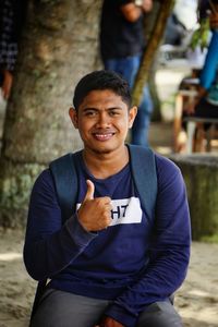 Portrait of smiling young man showing thumbs up against tree