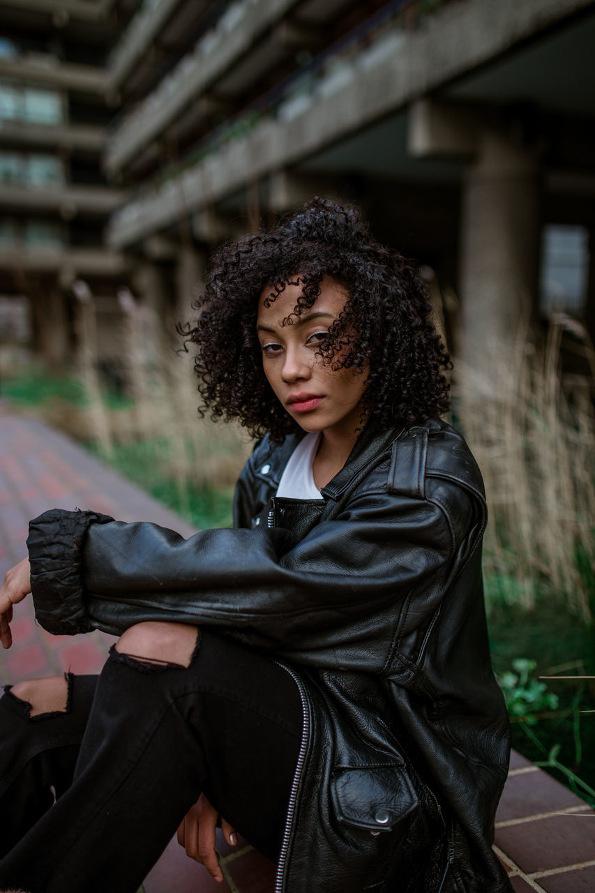 one person, sitting, young adult, lifestyles, young women, real people, focus on foreground, architecture, curly hair, portrait, adult, women, leisure activity, hairstyle, beauty, three quarter length, beautiful woman, clothing, jacket, hair, leather