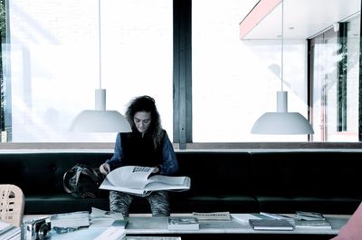 Young woman reading book while sitting on sofa against window