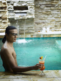 Shirtless young man holding drink while swimming in pool