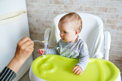 Cropped hand of mother feeding baby girl sitting on high chair against wall