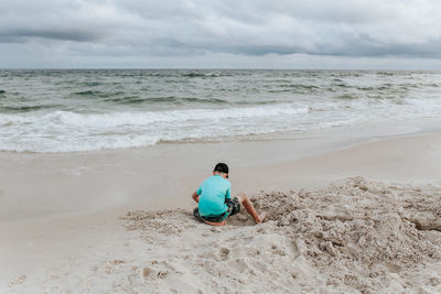 Rear view of man sitting on shore at beach against sky