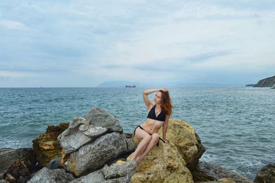 Young woman wearing bikini sitting on rock against sea and sky