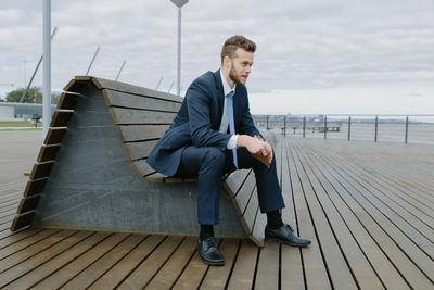Young man looking away while sitting on shore against sky