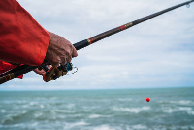 Hand holding fishing rod over sea against sky