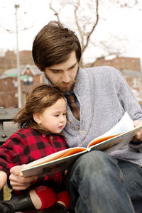 Father and daughter looking at book while sitting on bench in park