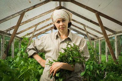 Portrait of woman holding plant while standing in greenhouse