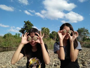 Female friends holding pebbles while standing at lakeshore against blue sky